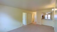 This single-floor residence offers a straightforward and functional layout centered around a spacious living area finished with wall-to-wall carpet in a neutral beige tone. The walls are painted in a light cream color, contributing to a bright and airy atmosphere throughout the space. Standard ceiling heights lend the room a comfortable, practical feel.

Along the back wall of the living area, a set of bifold louvered closet doors provides generous storage. Adjacent to the closet, an open doorway leads to a dedicated laundry area, where a stacked washer and dryer unit is visible, offering the convenience of in-unit laundry. A standard door nearby provides additional access to a separate room or hallway.

To the right side of the living area, a pass-through counter connects to the kitchen, allowing for an open sightline between the two spaces. The kitchen area, partially visible from this vantage point, features warm-toned countertops and accessible cabinetry. The overall layout is designed with practicality in mind, making efficient use of the available space while maintaining a clean, neutral aesthetic throughout.