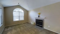 This single-story space centers around a welcoming living area distinguished by vaulted ceilings that lend a sense of openness and volume to the room. A prominent arched window on one wall allows abundant natural light to filter in, illuminating the neutral beige walls and carpeted flooring beneath. The vaulted ceiling angles gracefully upward, enhancing the airy quality of the space.

A tile-surround fireplace serves as a focal point, featuring a clean white mantel and a classic dark insert with brass accents. The hearth extends outward with coordinating tile work, grounding the fireplace as a functional and decorative centerpiece. The neutral tones of the walls and carpet create a versatile backdrop suited to a variety of interior styles.

The overall ambiance is calm and understated, with the combination of natural light from the arched window, the warmth suggested by the fireplace, and the open ceiling height contributing to a comfortable and inviting environment. The space is straightforward in its layout, offering a practical and well-lit living area with timeless architectural touches.