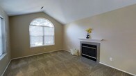 This single-story space centers around a welcoming living area distinguished by vaulted ceilings that lend a sense of openness and volume to the room. A prominent arched window on one wall allows abundant natural light to filter in, illuminating the neutral beige walls and carpeted flooring beneath. The vaulted ceiling angles gracefully upward, enhancing the airy quality of the space.

A tile-surround fireplace serves as a focal point, featuring a clean white mantel and a classic dark insert with brass accents. The hearth extends outward with coordinating tile work, grounding the fireplace as a functional and decorative centerpiece. The neutral tones of the walls and carpet create a versatile backdrop suited to a variety of interior styles.

The overall ambiance is calm and understated, with the combination of natural light from the arched window, the warmth suggested by the fireplace, and the open ceiling height contributing to a comfortable and inviting environment. The space is straightforward in its layout, offering a practical and well-lit living area with timeless architectural touches.