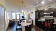 This single-floor space showcases a thoughtfully designed open-concept layout that seamlessly connects the kitchen, dining, and living areas into one cohesive and functional environment. The kitchen is a standout feature, equipped with rich dark cabinetry, granite countertops, and a breakfast bar with seating — ideal for casual dining or entertaining. Pendant lighting hangs above the bar, adding a warm, stylish focal point, while track lighting along the ceiling ensures the kitchen remains well-lit and practical. The dark hardwood flooring runs throughout, providing a consistent and elegant foundation that ties the entire space together. The dining area sits adjacent to the kitchen, offering a dedicated space for meals without feeling separated from the rest of the home. Beyond, the living area benefits from multiple windows that invite natural light, creating a bright and welcoming atmosphere. A ceiling fan in the living area adds comfort, and the neutral wall tones throughout lend a clean, versatile backdrop. The open flow between all areas makes the space feel expansive and connected, well-suited for both everyday living and hosting guests.