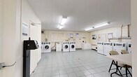 The laundry facility at 25 Seeley Dr is a practical, well-organized space spread across a single floor, comprising two dedicated laundry rooms. The facility is finished with light grey ceramic floor tiles throughout, offering a clean and durable surface that is easy to maintain. Walls are painted in neutral white tones with painted cinder block construction visible in areas, contributing to the utilitarian character of the space.

Each laundry room is equipped with multiple front-loading and top-loading washing machines alongside a row of dryers, all card-operated for convenient use. Bright overhead fluorescent lighting ensures the rooms are well-lit at all times. Ventilation ductwork is visible along the upper walls of the dryer area, supporting proper airflow and safety.

Additional amenities include a white utility counter with cabinet storage, a folding table with chairs for sorting laundry, and informational notices posted on the walls detailing machine usage guidelines. A utility sink is also present, adding further functionality to the space.

Access between the two rooms is provided through interior doors, and the layout allows for comfortable circulation even when machines are in use. The overall environment is clean, bright, and efficiently designed to serve the practical laundry needs of residents.