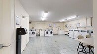 The laundry facility at 25 Seeley Dr is a practical, well-organized space spread across a single floor, comprising two dedicated laundry rooms. The facility is finished with light grey ceramic floor tiles throughout, offering a clean and durable surface that is easy to maintain. Walls are painted in neutral white tones with painted cinder block construction visible in areas, contributing to the utilitarian character of the space.

Each laundry room is equipped with multiple front-loading and top-loading washing machines alongside a row of dryers, all card-operated for convenient use. Bright overhead fluorescent lighting ensures the rooms are well-lit at all times. Ventilation ductwork is visible along the upper walls of the dryer area, supporting proper airflow and safety.

Additional amenities include a white utility counter with cabinet storage, a folding table with chairs for sorting laundry, and informational notices posted on the walls detailing machine usage guidelines. A utility sink is also present, adding further functionality to the space.

Access between the two rooms is provided through interior doors, and the layout allows for comfortable circulation even when machines are in use. The overall environment is clean, bright, and efficiently designed to serve the practical laundry needs of residents.