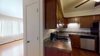 This single-floor residence offers a practical and cohesive layout designed for comfortable everyday living. The kitchen is a standout feature, showcasing rich, dark-toned wood cabinetry that extends to the upper walls, providing generous storage. Dark granite-style countertops complement the cabinetry, while a stainless steel electric range, stainless dishwasher, and a double-basin sink complete the functional workspace. Under-cabinet lighting adds a warm, practical touch to the cooking area. A decorative tile backsplash accent adds subtle character between the counters and upper cabinets. Warm hardwood flooring flows consistently throughout the space, lending a cohesive and inviting atmosphere. The living area is open and well-proportioned, bathed in natural light from large windows fitted with vertical blinds. A wall-mounted air conditioning unit and baseboard heating ensure year-round comfort. The bathroom features a full soaking tub with a shower, a wood-toned vanity with a white countertop, and a large mirror, all tied together with matching hardwood flooring. The overall layout, as revealed by the floorplan view, is efficiently organized, connecting the main living area, kitchen, and bathroom in a straightforward and accessible arrangement.