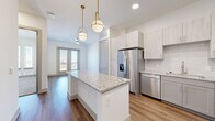Located at 10221 Whistle Stop Lane in Frisco, TX, this thoughtfully designed one-bedroom, one-bathroom residence offers a clean, modern aesthetic across a single floor. The open-concept kitchen and living area serves as the heart of the home, showcasing a granite-topped central island illuminated by stylish gold pendant lights. The kitchen is equipped with stainless steel appliances—including a French door refrigerator, range, microwave, and dishwasher—complemented by two-tone cabinetry in light and gray finishes, a subway tile backsplash, and wood-look flooring. A dedicated pantry provides additional storage. The living area flows naturally toward glass double doors that open onto a private balcony, allowing ample natural light to fill the space. The bedroom features plush carpet flooring, large windows with outdoor views, and a ceiling fan. It connects directly to the full bathroom, which includes a tiled soaking tub/shower combo, granite vanity, and wood-look flooring, with a walk-in closet conveniently accessible from within. A dedicated laundry area rounds out the functional layout of this well-appointed Frisco residence.