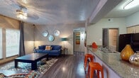 This single-floor residence offers a thoughtfully designed open-concept layout that connects the living, dining, and kitchen areas into a cohesive, functional space. The living room features wood-style flooring, a textured ceiling, and large windows that admit generous natural light. A defined dining area accommodates a round table and seating, with neutral walls providing a clean backdrop.

The kitchen is equipped with light wood cabinetry, granite-style countertops, a tile backsplash, and black appliances including a refrigerator, electric range, and microwave. A peninsula counter with bar seating connects the kitchen directly to the living area, enhancing the open feel of the space.

The bedroom is carpeted and includes a ceiling fan, a window with outdoor views, and ample room for full-sized furniture. The bathroom features wood-style flooring, a vanity with granite-style countertops, light wood cabinetry, a large mirror, and a shower-tub combination. Throughout the unit, a consistent warm and neutral color palette is complemented by contemporary accent tones, creating a cohesive and inviting atmosphere.
