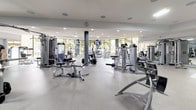 This expansive fitness center is designed to accommodate a full range of training needs within a single, open-plan floor. The space features a clean, contemporary aesthetic characterized by white and grey tones, smooth speckled flooring, and a well-lit environment created by a combination of recessed ceiling lights and large floor-to-ceiling windows. These windows frame views of tropical palm trees, allowing ample natural light to fill the facility and contributing to an open, airy atmosphere.

The strength training area is extensively equipped with a wide array of Cybex selectorized machines targeting major muscle groups, a dedicated free weights section with multiple weight plate racks and adjustable benches, and a cable machine zone. Large mirrors line one section of the wall, aiding form checks during exercise.

A functional training area features TRX suspension trainers, medicine balls, stability balls, and resistance tools organized along a mirrored wall. A reception or check-in station with computer terminals is centrally positioned, providing staff oversight of the floor. The facility's curved architectural elements, including rounded ceiling details and cylindrical support columns, add a refined design quality to the overall space.