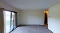 This single-floor residential space offers a clean, straightforward layout designed for comfortable everyday living. The main living area is finished with wall-to-wall carpeting that adds warmth underfoot, while smooth white walls and a bright ceiling create a neutral, move-in-ready canvas. A large sliding glass door dominates one side of the room, flooding the space with natural light and providing a direct view of the exterior brick facade and outdoor area beyond — a functional feature that also enhances the sense of openness. Along the base of the far wall, a white baseboard heating unit runs the length of the room, ensuring efficient and even heat distribution throughout. Electrical outlets are conveniently positioned along the walls. A hallway visible from the living area leads to additional rooms, suggesting a well-connected interior layout. The overall ambiance is practical and understated, with a simple, uncluttered design that allows the space to be easily personalized. The single-floor configuration makes for seamless, step-free navigation throughout the entire unit.