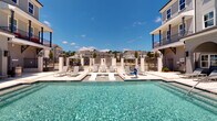 This amenity space features two distinct areas designed to support an active lifestyle. The outdoor pool area is a standout feature, showcasing a generously sized swimming pool with crystal-clear water, flanked by a broad paved deck furnished with lounge chairs and market umbrellas. A built-in spa or wading feature sits adjacent to the main pool, and outdoor grilling stations are available nearby. The surrounding fenced enclosure provides privacy while maintaining views of the neighboring community and lush landscaping under open skies.

The fitness center is thoughtfully laid out across a well-lit interior space, featuring herringbone-patterned carpet flooring and soaring ceilings with recessed lighting and ceiling fans. Large windows along multiple walls flood the space with natural light and offer views of the outdoors. The facility is comprehensively equipped with Precor cardio machines, treadmills, a rowing machine, a multi-functional cable and power rack system, a full dumbbell rack, medicine balls, battle ropes, and stability balls. Floor-to-ceiling mirrors along one wall enhance the sense of space. A wall-mounted television in the cardio area adds convenience for workout entertainment.