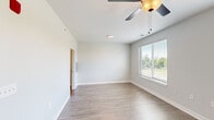 This single-floor residence features a clean, contemporary design with a neutral color palette and wood-look flooring that flows consistently throughout the space. The open-concept layout connects the living area to the kitchen, creating a cohesive and functional floor plan.

The living area is well-lit by multiple windows fitted with blinds, allowing ample natural light while maintaining privacy. A ceiling fan with a gold-accented fixture adds a stylish touch overhead. The soft gray walls and crisp white trim contribute to a bright, modern aesthetic.

The kitchen is a standout feature, offering rich dark-toned cabinetry with brushed nickel hardware, granite countertops, and a classic white subway tile backsplash. A large center island with a built-in sink and pendant lighting serves as both a functional workspace and a gathering point. Stainless steel appliances, including a refrigerator, range, and over-the-range microwave, complete the kitchen.

The bathroom features the same wood-look flooring, a frosted glass shower enclosure, and a clean, minimalist design with white walls and trim. The space overall presents a well-appointed, move-in-ready environment with modern finishes throughout.