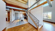 This two-story loft-style space showcases a distinctive blend of industrial and rustic design elements throughout. The main level features warm hardwood flooring and an open layout that connects the living area to a compact kitchen. The kitchen is equipped with stainless steel appliances, including a refrigerator, range, and microwave, complemented by rich dark wood cabinetry and tile flooring. A wood-framed staircase with black metal balusters leads to the upper mezzanine level, which is finished with plush carpet and bordered by an open railing that overlooks the main floor below.

Throughout both levels, exposed heavy timber posts and beams contribute to the space's structural character, while tongue-and-groove wood plank ceilings add warmth overhead. Visible HVAC ductwork runs along the ceiling, reinforcing the industrial aesthetic. Windows on the upper level allow natural light to filter in and offer views of the surrounding neighborhood. A full bathroom on the lower level includes a walk-in shower and tile flooring. Additional rooms on the upper level feature carpeted floors, exposed beam ceilings, and windows providing natural light, creating versatile spaces suitable for a variety of uses.
