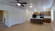 This single-floor residence offers a thoughtfully designed open-concept layout that effortlessly connects the living and kitchen areas, creating a sense of spaciousness and flow. The kitchen is a standout feature, showcasing rich wood-tone cabinetry, a sleek kitchen island with a light countertop and built-in sink, and pendant lighting that adds a warm, contemporary touch. Black appliances, including a refrigerator and range, complement the modern aesthetic while offering full functionality. The living area is finished with light-toned flooring that enhances the bright, airy atmosphere throughout the space. A ceiling fan with an integrated light fixture provides both comfort and illumination, while additional flush-mount ceiling lights ensure consistent, well-distributed lighting across the open floor plan. Crisp white walls and clean architectural lines give the interior a fresh, minimalist character. Paneled white doors lead to additional rooms or storage areas, maintaining the cohesive and uncluttered design language. Overall, the space presents a clean, modern aesthetic that balances style and practicality in a well-appointed, move-in-ready layout.
