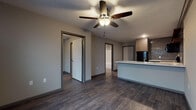 Located at 1435 W Talmage St in Springfield, Missouri, this single-floor unit offers a cohesive, modern aesthetic throughout. Dark hardwood-style flooring runs continuously across the living areas, creating a unified and contemporary feel.

The open living area is anchored by a ceiling fan with integrated lighting and connects seamlessly to the kitchen via a peninsula-style counter with a light stone surface, ideal for casual dining or meal prep. The kitchen is well-appointed with wood-toned lower and upper cabinetry, open shelving with decorative tile backsplash, and a full suite of black appliances including a refrigerator, range, microwave, and ample counter space.

The bedroom is a comfortably sized, private room outfitted with a ceiling fan and access to an adjacent laundry area with washer and dryer hookups, adding significant convenience.

The bathroom features a dark wood vanity with a marble-style countertop, a large mirror with overhead lighting, and a full-size soaking tub and shower combination with clean white surrounds and hardwood-style flooring extending throughout.

Neutral gray walls and dark trim accents tie the space together, lending a clean, modern sensibility throughout. The layout is efficient and well-organized, making this unit a practical and stylish option in Springfield, MO.