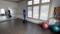 This single-floor fitness studio is thoughtfully designed to support a variety of exercise routines and training activities. The space features durable dark hardwood-style flooring that provides both resilience and a clean, professional aesthetic suited for high-impact workouts. A full-length mirrored wall lines one side of the room, allowing users to monitor their form and technique during exercise. The studio is bathed in natural light courtesy of large, multi-pane windows fitted with adjustable blinds, offering views of the surrounding greenery while maintaining privacy as needed. The elevated transom windows above further enhance the brightness of the space. Crown molding and neutral-toned walls contribute a polished, well-finished look to the room. Equipment such as a freestanding punching bag and an exercise ball are present, illustrating the space's versatility for both cardio and strength training. The open layout provides ample room for stretching, yoga, aerobics, or martial arts practice. Overall, this fitness studio combines functional design with inviting architectural details, making it a well-rounded space for health and wellness activities.