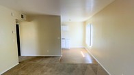 This single-story space offers a straightforward, functional layout designed for comfortable everyday living. The main living area is finished with wall-to-wall carpeting, providing a soft, warm underfoot feel, while the adjoining kitchen and transitional areas feature smooth hardwood-style flooring that adds a clean contrast. The open sightlines between the living space and kitchen create a connected, airy feel throughout.

The kitchen is positioned just beyond a partial dividing wall, which adds subtle definition between the spaces without fully closing them off. It features white cabinetry that runs along the lower portion of the walls, complemented by a sink and ample counter space, keeping the area bright and practical. A wall-mounted cabinet above provides additional storage.

The living area benefits from a large window fitted with vertical blinds, allowing natural light to filter in and brighten the neutral, cream-toned walls. The overall color palette throughout the space is light and consistent, contributing to a clean, move-in ready aesthetic. The single-floor layout ensures easy accessibility and seamless flow between the living and kitchen areas.