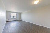 Located on the 14th floor at 940 Commissioners Rd in London, Ontario, this one-bedroom, one-bathroom unit offers a clean, modern aesthetic with a practical and efficient layout. The living room is a bright, open space featuring light grey laminate flooring, neutral white walls, and a large window that frames expansive city views — flooding the room with natural light and offering a connection to the London skyline. A baseboard heating unit runs beneath the window, ensuring year-round comfort. The bedroom continues the same laminate flooring and neutral palette, with its own window providing additional natural light and elevated views of the surrounding cityscape. The kitchen is compact and functional, featuring dark granite-style countertops, white cabinetry, and a white electric range — offering everything needed for day-to-day meal preparation. The bathroom is well-appointed with a full tub-shower combo, a white vanity topped with a dark granite-style counter, a large mirror with overhead lighting, and tile flooring. Throughout the unit, the consistently neutral tones and clean finishes create a calm, move-in-ready atmosphere suited for comfortable urban living.