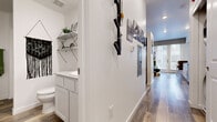 This single-floor unit offers a cohesive modern design throughout, unified by warm wood-look plank flooring and crisp white walls that create a clean, inviting atmosphere. The living area serves as the heart of the space, featuring recessed lighting, a large window that welcomes natural light, and built-in shelving that adds both function and character. The room comfortably accommodates a sectional sofa arrangement and includes a dedicated workspace nook, making it well-suited for both relaxing and working from home. The kitchen is thoughtfully outfitted with white shaker-style cabinetry, granite-look countertops, a stainless steel dishwasher, and a full range with overhead microwave, providing everything needed for everyday cooking. A hallway connects the main living spaces and is accented with framed wall art, a wall-mounted organizer, and a thermostat panel. The bathroom features a white vanity with ample cabinet storage, geometric open shelving, and decorative touches including a macramé wall hanging and framed artwork, lending a curated, boutique feel. The overall ambiance is contemporary and well-balanced, blending practical functionality with thoughtful design details throughout.