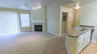 This single-floor residence offers a thoughtfully designed layout with an open-concept living area that immediately draws the eye with its impressive vaulted ceilings. A ceiling fan with integrated lighting anchors the space, enhancing airflow and ambiance. The living room is centered around a classic white-mantled fireplace with elegant glass doors, serving as a natural focal point and lending warmth to the room. Plush wall-to-wall carpeting in a neutral tone runs throughout the living area, contributing to a comfortable and inviting atmosphere. Natural light filters in through a window fitted with blinds, brightening the soft, neutral-toned walls. To one side, a curved kitchen bar counter with a granite-style surface transitions smoothly into the kitchen, which features a sink and overhead lighting — ideal for both casual dining and entertaining. A hallway visible from the main living space leads to additional rooms, including what appears to be a bathroom and a secondary room, suggesting a practical and well-organized floor plan. The overall aesthetic is clean, neutral, and move-in ready, offering a versatile canvas for a variety of interior styles.