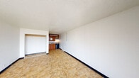 This single-floor unit offers a straightforward, open layout that combines the living area and sleeping space into one continuous room. The main living area is finished with tile flooring in a warm earth tone and features white walls accented by dark baseboards, creating a clean, neutral canvas. A textured popcorn ceiling runs throughout the unit. An open closet with a hanging rod is positioned along one wall, providing convenient storage without the need for a separate room.

The kitchenette is set into a defined alcove and includes warm-toned wood cabinetry both above and below the counter, a single-basin sink with a stone-tile backsplash, a compact cooktop with an overhead range hood, and a black mini-refrigerator. The kitchen area is well-lit by an under-cabinet light fixture.

At the far end of the main room, a large sliding window paired with an entry door allows natural light to fill the space and offers views of the surrounding outdoor environment. A wall-mounted HVAC unit positioned beneath the window provides climate control.

The bathroom is compact and fully tiled in a classic light pink ceramic tile, featuring a full-size soaking tub with an overhead shower, a wall-mounted pedestal sink, and a toilet. A wall-mounted mirror and overhead lighting complete the bathroom's functional design.