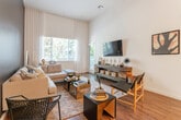 This well-appointed single-story residence showcases a thoughtfully designed open-concept layout that seamlessly connects the kitchen and living areas. The kitchen is a standout feature, outfitted with stainless steel appliances including a French door refrigerator, gas range, built-in microwave, and dishwasher. Two-toned cabinetry — crisp white uppers paired with dark lower cabinets — frames the space, while a clean white countertop peninsula provides additional prep and casual dining space. Pendant lighting overhead adds a modern finishing touch. The living room impresses with dramatically soaring ceilings and large windows that invite an abundance of natural light and views of surrounding greenery. A bold teal accent wall anchors the living area, complementing the rich hardwood-style flooring that runs throughout the main spaces. The bedroom is a serene retreat, featuring the same elevated ceilings, plush carpeting, tall windows with sheer drapery, and direct access to both a walk-in closet and a private en-suite bathroom. The bathroom includes a dual-sink vanity with stone-look countertops and a full soaking tub with subway tile surround. The overall ambiance is contemporary and inviting, with a sophisticated palette of neutral tones accented by deep, rich colors.