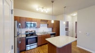 This single-floor residence is centered around a thoughtfully designed kitchen that serves as the focal point of the living space. The kitchen is outfitted with a full suite of stainless steel appliances, including a range, microwave, refrigerator, and dishwasher. Rich, warm-toned cabinetry lines the walls, complemented by granite countertops that extend to a freestanding island, providing additional prep space and a casual dining option. A decorative lattice cabinet detail adds visual interest above the main counter run. Pendant lighting hangs above the island, while a track lighting fixture illuminates the primary cooking and prep areas, creating a well-lit, functional environment. Hardwood-style flooring runs throughout the kitchen and extends into the open living area, which is spacious and bright, featuring a ceiling fan and a door leading to an adjoining carpeted room. Natural light filters in through windows along the living area's exterior wall. The bathroom includes a soaking tub with shower, hardwood-style flooring consistent with the rest of the unit, granite-topped vanity, and clean, neutral finishes throughout.