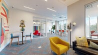 This dynamic, single-floor workspace is thoughtfully designed to balance open collaboration with private meeting areas. The layout features a generous open-plan lounge zone anchored by bold, mid-century modern furnishings — including a mustard yellow armchair, a dark leather sofa, and accent chairs in red, teal, and blue — creating a visually stimulating yet functional environment. A herringbone-patterned carpet in warm grey with striking teal accents runs throughout, unifying the space with a contemporary aesthetic.

Glass-walled meeting rooms with sliding doors provide semi-private settings for focused gatherings, while remaining visually connected to the broader workspace. Inside these rooms, high-top bar tables and colorful seating continue the space's vibrant design language. A separate lounge nook, defined by a vertical slat partition, offers a more intimate setting with a leather sofa and accent chair.

Floor-to-ceiling windows line the perimeter, inviting abundant natural light and offering open views of the surrounding cityscape and greenery. Recessed linear lighting and pendant fixtures supplement the natural illumination, ensuring a well-lit environment throughout the day. Decorative indoor plants and curated art pieces add warmth and personality, completing this energetic, design-forward workspace.