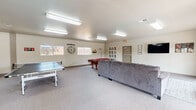 This single-story community clubhouse is designed to serve as a versatile gathering space, offering a range of amenities across its open, carpeted interior. The game room is a central highlight, featuring a classic red felt pool table, a ping-pong table, and a large sectional sofa positioned in front of a wall-mounted flat-screen TV. Playful wall décor, including oversized XO lettering and a statement clock, adds character to the bright, neutral-toned walls illuminated by fluorescent overhead lighting.

Adjacent to the game room, a lounge area benefits from abundant natural light streaming through large French doors, which open directly onto an outdoor patio and pool area, providing a seamless indoor-outdoor connection. The dining area accommodates a sizable wooden table with chairs and a padded bench, suitable for group gatherings or meetings. Nearby, a compact kitchenette is equipped with wood-toned cabinetry, a refrigerator, microwave, and a small countertop bar with bar stools, making the space practical for catered events or casual use. Throughout, the consistent light carpet, neutral wall colors, and functional furnishings create a clean, welcoming atmosphere suited for community activities.