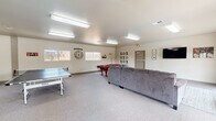 This single-story community clubhouse is designed to serve as a versatile gathering space, offering a range of amenities across its open, carpeted interior. The game room is a central highlight, featuring a classic red felt pool table, a ping-pong table, and a large sectional sofa positioned in front of a wall-mounted flat-screen TV. Playful wall décor, including oversized XO lettering and a statement clock, adds character to the bright, neutral-toned walls illuminated by fluorescent overhead lighting.

Adjacent to the game room, a lounge area benefits from abundant natural light streaming through large French doors, which open directly onto an outdoor patio and pool area, providing a seamless indoor-outdoor connection. The dining area accommodates a sizable wooden table with chairs and a padded bench, suitable for group gatherings or meetings. Nearby, a compact kitchenette is equipped with wood-toned cabinetry, a refrigerator, microwave, and a small countertop bar with bar stools, making the space practical for catered events or casual use. Throughout, the consistent light carpet, neutral wall colors, and functional furnishings create a clean, welcoming atmosphere suited for community activities.