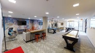 This single-floor entertainment space is thoughtfully designed to deliver a vibrant social experience through a blend of industrial and modern aesthetics. A striking exposed brick accent wall, adorned with colorful mural artwork, serves as a dramatic focal point and anchors a bar-height counter complete with a wooden top and red metal barstools — perfect for casual gathering and conversation. A wall-mounted flat-screen television completes the lounge vibe in this area. Moving deeper into the space, a full-sized pool table takes center stage beneath an eye-catching arrangement of decorative pendant chandeliers, adding both elegance and character to the room. A shuffleboard table and additional game surfaces invite friendly competition and extended play. A high-top bar counter with yellow barstools defines a secondary social zone, providing additional seating and versatility. Large windows along the perimeter flood the space with natural light, enhancing the open, airy feel of the room. Recessed ceiling lighting complements the ambient glow throughout. The flooring features a patterned carpet that adds warmth while tying together the eclectic design elements. Overall, the space strikes a confident balance between energetic entertainment and refined style.