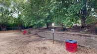 This outdoor dog park offers a spacious, fully enclosed area designed for off-leash canine activity and recreation. The ground is covered with a generous layer of wood chip mulch, providing a comfortable and low-maintenance surface for dogs of all sizes to run and play freely.

The park is secured by a combination of chain-link fencing along the back and sides, and a classic white picket fence along one border, ensuring safe containment. Mature trees line the perimeter, offering natural shade and a lush, green backdrop that enhances the park's welcoming atmosphere.

Several agility and play elements are scattered throughout the space, including a blue barrel tunnel, hurdle bars, tire obstacles, and a mock fire hydrant, providing mental and physical stimulation for visiting dogs. A raised retaining wall with landscaped plantings adds visual interest and a defined boundary to one side of the park.

Practical amenities are thoughtfully placed throughout, including a pet waste bag dispenser station and a prominently placed red waste bin, encouraging responsible pet ownership. A red bench is also available, offering a comfortable spot for pet owners to sit and supervise their dogs. The open sky above and surrounding greenery create a bright, airy environment that makes this dog park both functional and enjoyable for pets and their owners alike.