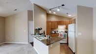 This single-floor residence offers a well-designed open-concept layout that seamlessly connects the kitchen and living areas. The kitchen is a standout feature, showcasing warm honey-toned wood cabinetry that provides generous storage, complemented by rich dark granite countertops. A peninsula-style breakfast bar extends the counter space and offers a casual dining area, making the kitchen both functional and inviting for everyday living and entertaining.

The kitchen is equipped with white appliances, including a refrigerator, range, and overhead ventilation hood. Track lighting mounted along the vaulted ceiling provides directed task illumination, while recessed lighting adds a warm ambient glow throughout the space. The soaring vaulted ceilings amplify the sense of openness and airiness, contributing to a spacious feel.

Neutral warm beige tones on the walls create a cohesive and welcoming atmosphere throughout the space. The flooring transitions from durable hardwood-style planks in the kitchen area to plush carpeting in the adjoining living area, adding warmth and comfort underfoot. This thoughtfully laid-out residence balances practicality with a comfortable, homey aesthetic that suits a variety of lifestyles.