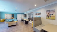 Located at 1347 Dorado Dr in Tallahassee, FL, this single-floor community game and social lounge is designed to encourage recreation and social interaction. The space is finished with light wood-look flooring and accented by teal and soft blue walls, creating an energetic yet welcoming atmosphere throughout.

The main game area serves as the focal point of the lounge, housing a full-sized pool table with blue felt and a regulation shuffleboard table. A large wall-mounted flat-screen television is positioned nearby, adding to the entertainment options. A oversized framed Scrabble board mounted on the wall adds a playful, decorative touch, while mustard yellow accent chairs provide comfortable casual seating in this area.

A dedicated lounge nook features high-back upholstered booth seating arranged around a pedestal table, offering a more intimate gathering spot. The walls are adorned with colorful local and collegiate-themed artwork, giving the space a community-oriented character.

A bright café-style seating area with large windows allows abundant natural light to pour in, offering outdoor views of the surrounding greenery. This section features white high-top tables paired with teal metal chairs and contemporary pendant lighting. Recessed lighting throughout ensures the entire space remains well-lit, completing a lively and functional social environment.