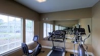 This single-floor fitness room is designed to support an active lifestyle, offering a focused and functional workout environment. The space is outfitted with a selection of cardio equipment, including a treadmill and a stationary recumbent bike, making it suitable for a variety of exercise routines. A large floor-to-ceiling mirror spans one wall, visually expanding the room and allowing users to monitor their form during workouts. The warm, earthy-toned walls create a grounded, focused atmosphere, while the carpeted flooring adds a layer of comfort underfoot. Multiple windows fitted with white horizontal blinds line the walls, inviting ample natural light and offering views of surrounding greenery, contributing to an open and energizing ambiance. The ceiling features recessed lighting to ensure the space remains well-lit at all times of day. The layout is thoughtfully arranged to allow for ease of movement between machines. Overall, this fitness room combines practical equipment, smart design, and a welcoming atmosphere to provide an effective and comfortable space for daily exercise.