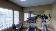 This single-floor fitness room is designed to support an active lifestyle, offering a focused and functional workout environment. The space is outfitted with a selection of cardio equipment, including a treadmill and a stationary recumbent bike, making it suitable for a variety of exercise routines. A large floor-to-ceiling mirror spans one wall, visually expanding the room and allowing users to monitor their form during workouts. The warm, earthy-toned walls create a grounded, focused atmosphere, while the carpeted flooring adds a layer of comfort underfoot. Multiple windows fitted with white horizontal blinds line the walls, inviting ample natural light and offering views of surrounding greenery, contributing to an open and energizing ambiance. The ceiling features recessed lighting to ensure the space remains well-lit at all times of day. The layout is thoughtfully arranged to allow for ease of movement between machines. Overall, this fitness room combines practical equipment, smart design, and a welcoming atmosphere to provide an effective and comfortable space for daily exercise.