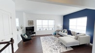 This well-appointed two-floor residence combines contemporary style with comfortable living across a thoughtfully designed layout. The living area immediately captures attention with its striking navy blue accent wall, which provides a bold contrast against the crisp white walls and ceiling. Rich, dark hardwood flooring runs throughout, lending warmth and continuity to the space. Natural light pours in through multiple windows dressed with clean white roller shades, creating a bright and airy atmosphere. The living room is generously proportioned, comfortably accommodating a large sectional sofa, an accent chair, and a media console, all anchored by an intricately patterned area rug in soft grey and white tones. A sleek flat-screen television serves as a focal point on one wall, while decorative accents in teal and navy echo the home's cohesive color palette. The open layout promotes an easy flow between living and entertaining areas. Subtle architectural details, including ceiling beams and clean-lined trim work, add depth and character throughout. The overall ambiance strikes a balance between modern sophistication and everyday comfort, making this a space that is both visually engaging and highly functional across its two levels.