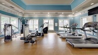 Located at 465 Elmcroft Blvd in Rockville, Maryland, this well-appointed fitness center offers a comprehensive workout environment within a single, open floor plan. The space is distinguished by its elegant teal-painted walls, crisp white crown molding, and a decorative tray ceiling with recessed lighting, creating a bright and upscale atmosphere. Cork-style flooring provides a durable yet comfortable surface throughout the workout area. Large white-framed windows along multiple walls allow abundant natural light to fill the room, while strategically placed mirrors amplify the sense of space and support exercise form. The cardio section is well-equipped with multiple treadmills, elliptical trainers, and a stair climber, each positioned to take advantage of the natural views outside. The strength training area includes a multi-station cable machine, an adjustable weight bench, and a full rack of dumbbells spanning a wide range of weights. Fitness accessories such as stability balls, resistance bands, and foam rollers are neatly organized on dedicated racks. Wall-mounted televisions provide entertainment during cardio sessions, and a hand sanitizer station ensures a clean environment. A set of bold blue double doors serves as the main entrance, completing the cohesive and polished design of this community fitness facility.