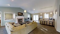 This single-floor communal space is thoughtfully designed to provide a comfortable and functional environment for gathering and relaxation. The main lounge area is anchored by a classic white-framed fireplace, above which a wall-mounted flat-screen TV is positioned as a central focal point. Plush, generously sized seating in warm golden tones is arranged around a dark wood coffee table, creating an inviting area for conversation and leisure.

The vaulted ceilings rise dramatically overhead, enhancing the sense of openness and airiness throughout the space. Soft sage-green walls contribute a calm, neutral palette that complements the natural light streaming in through multiple windows and a set of French doors, which connect the interior to the outdoors.

Stylish pendant lighting — including a globe-shaped fixture over the lounge and sleek bar pendants — adds a modern touch to the overall aesthetic. Toward one end of the space, a café-style bar counter with dark cabinetry and bar stool seating provides a casual spot for refreshments, complete with a coffee station. An additional high-top table and chairs offer a more upright seating alternative. The combination of warm tones, natural light, and varied seating arrangements makes this a versatile and welcoming communal retreat.