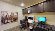 This single-floor business center is thoughtfully designed to provide a functional and efficient workspace. The room features a continuous built-in countertop workstation that spans the walls, offering ample desk space for multiple users simultaneously. Above the workstation, a row of dark espresso-finished cabinetry provides generous closed storage, keeping the area organized and professional in appearance. The space is equipped with desktop computers and a multifunction printer/scanner/copier, making it well-suited for a variety of business tasks. Ergonomic office chairs accompany each workstation for comfortable use. The flooring features a bold patterned carpet that adds a touch of character to the otherwise neutral interior. Warm beige walls and recessed ceiling lighting create a bright and welcoming atmosphere, while a piece of decorative wall art adds a finishing touch. The textured ceiling complements the overall design aesthetic. Whether used for printing, scanning, or general computer work, this business center is a practical and accessible resource for guests or residents seeking a dedicated workspace.