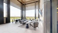Located at Whistle Stop Lane in Frisco, TX, this contemporary rooftop lounge seamlessly blends indoor comfort with open-air living. The space is defined by soaring ceilings, large-format light-toned tile flooring, and a striking architectural pergola structure that frames panoramic views of the Frisco skyline and surrounding greenery.

The interior living area is anchored by a sleek linear fireplace set into a smooth, neutral-toned accent wall, above which a wall-mounted flat-screen TV is positioned for comfortable viewing. Stylish woven-frame lounge chairs and accent tables are arranged atop a geometric area rug, creating an inviting gathering atmosphere. Tall windows and glass doors flood the space with abundant natural light.

Transitioning outdoors, the expansive terrace offers multiple seating configurations, including dining tables with coordinating chairs suited for group gatherings, as well as casual lounge seating. Dark architectural columns punctuate the open-air deck, providing structural elegance while supporting the overhead slatted pergola that filters sunlight throughout the day. Glass railings ensure unobstructed views of the Frisco cityscape and open green spaces beyond. The terrace also features convenient access points through multiple entry doors, making the space highly functional for community use.