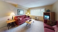 This single-floor residence at 501 Plantation St in Worcester, MA offers a thoughtfully designed layout with a warm, neutral color palette throughout. The living area features plush carpeting, a comfortable seating arrangement, and wall art that adds a welcoming touch, with natural light filtering through vertical blinds. The kitchen is well-equipped with rich wood cabinetry, granite-style countertops, black appliances including an electric range, and wood-look flooring, providing an efficient and functional cooking space. The primary bedroom is generously sized, featuring carpet flooring, a metal-frame bed, a nightstand with lamp, a seating area, and access to a walk-in closet with wire shelving for ample storage. A second bedroom offers flexible use with carpeted floors, multiple doors leading to closet and bathroom access, and a relaxed, comfortable ambiance. Both bathrooms are finished with wood-look tile flooring, rich wood vanities with white countertops, large mirrors, and full bathtub-shower combinations. A dedicated sitting room with a plush white armchair and neutral décor provides a quiet retreat. The overall design emphasizes functional comfort with consistent warm tones and practical finishes throughout.