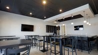 This single-floor space offers a contemporary, open-concept layout combining a lounge and kitchenette area. The interior is defined by a striking black drop-tile ceiling with recessed lighting and pendant fixtures that cast warm, focused light throughout the room. Crisp white walls contrast sharply with the dark ceiling, creating a bold, modern aesthetic.

The seating area features multiple high-top tables paired with metal bar-height chairs with cushioned seats, accommodating a generous number of guests. Wall-mounted flat-screen televisions are positioned for easy viewing from multiple seating arrangements. Light-toned tile flooring runs throughout, adding brightness to balance the darker upper elements.

The integrated kitchenette at the rear is equipped with dark cabinetry, granite-style countertops, and stainless steel appliances, including a full-size refrigerator. Pendant lights hang above the counter area, enhancing both functionality and style. An arched doorway leads to an adjoining space. Overall, the room presents a polished, functional environment well-suited for social gatherings or community use.