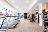 Located at 23 US-1 S in New Brunswick, NJ, this fitness center offers a thoroughly equipped workout facility designed to support a wide range of exercise routines. The space is organized into distinct functional zones, accommodating both strength training and cardiovascular exercise within a generously sized, open floor plan.

The strength training area features a robust lineup of Atlantis cable machines, selectorized resistance equipment, a multi-functional cable crossover station, and a leg press machine, providing comprehensive options for full-body training. Large wall-mounted mirrors throughout amplify the sense of space and allow users to monitor their form during workouts. A wall-mounted flat-screen television adds an entertainment element to the training environment.

The cardio zone is lined with treadmills and elliptical machines positioned near floor-to-ceiling windows, allowing natural light to fill the space and offering views of the exterior surroundings. Recessed lighting throughout the facility ensures consistent, even illumination across all areas.

Soft, neutral-toned carpet covers the flooring throughout, contributing to a clean, modern aesthetic. The overall design is contemporary and functional, with white walls, a tray ceiling detail, and a well-organized layout that promotes efficient use of the available space.