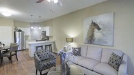 Located in McKinney, Texas, this single-floor residence offers a thoughtfully designed open-concept layout that seamlessly blends the living and kitchen areas into one cohesive, welcoming space. The living area is anchored by warm neutral walls that create a calm, inviting ambiance, complemented by wood-look flooring that adds warmth and continuity throughout. A ceiling fan overhead provides both comfort and a subtle architectural accent. The space is styled with comfortable seating, including a plush sofa and patterned accent chairs, lending the room a balanced mix of function and personality. A striking large-scale equestrian wall art piece serves as a bold focal point, adding character to the living area. The kitchen is equipped with white cabinetry, stainless steel appliances, and a mosaic tile backsplash that adds visual texture. A breakfast bar with seating bridges the kitchen and living areas, making it ideal for casual dining and entertaining. Pendant lighting above the bar adds a refined touch. Vaulted or angled ceilings contribute to an airy sense of volume and openness throughout the space. Overall, this residence delivers a clean, modern aesthetic with practical design choices suited for comfortable everyday living in McKinney, TX.