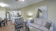Located in McKinney, Texas, this single-floor residence offers a thoughtfully designed open-concept layout that seamlessly blends the living and kitchen areas into one cohesive, welcoming space. The living area is anchored by warm neutral walls that create a calm, inviting ambiance, complemented by wood-look flooring that adds warmth and continuity throughout. A ceiling fan overhead provides both comfort and a subtle architectural accent. The space is styled with comfortable seating, including a plush sofa and patterned accent chairs, lending the room a balanced mix of function and personality. A striking large-scale equestrian wall art piece serves as a bold focal point, adding character to the living area. The kitchen is equipped with white cabinetry, stainless steel appliances, and a mosaic tile backsplash that adds visual texture. A breakfast bar with seating bridges the kitchen and living areas, making it ideal for casual dining and entertaining. Pendant lighting above the bar adds a refined touch. Vaulted or angled ceilings contribute to an airy sense of volume and openness throughout the space. Overall, this residence delivers a clean, modern aesthetic with practical design choices suited for comfortable everyday living in McKinney, TX.