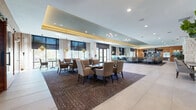 This expansive single-floor venue offers a sophisticated and versatile gathering space defined by refined architectural details and a warm, contemporary aesthetic. The open floor plan accommodates multiple seating configurations, from long communal dining tables to intimate clustered arrangements, all set atop richly patterned area rugs over large-format tile flooring. A coffered ceiling with integrated LED cove lighting in warm gold tones adds depth and a polished ambiance throughout the space. Floor-to-ceiling windows and multiple sets of glass double doors flood the interior with abundant natural light while offering views of the surrounding outdoor greenery. Distinctive design elements include a striking white stone accent wall that anchors the central corridor, warm wood-paneled walls, and oversized sliding barn-style doors that lend a transitional, upscale character. Pendant lighting in brushed gold and geometric finishes are suspended at varying heights, complementing the overall design palette of neutral tones, rich browns, and soft grays. A bar or service area with marble-finish countertops and a wine refrigerator is visible toward one end of the space, suggesting hospitality-forward functionality. The combination of open gathering areas, defined dining zones, and seamless indoor-outdoor connectivity makes this a highly functional and visually compelling event space.