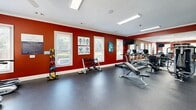 Located at 3606 Kaliste Saloom Rd in Lafayette, Louisiana, this dedicated fitness center offers a fully outfitted exercise environment designed to support a wide range of workout routines. The space is defined by bold, deep-red walls accented by crisp white crown molding and trim, creating an energetic and motivating atmosphere. The durable rubber flooring provides a safe and practical surface suited for both cardio and strength training activities. Multiple windows fitted with blinds allow ample natural light to fill the room, contributing to an open and airy feel. A large wall of floor-to-ceiling mirrors spans one side of the space, visually expanding the room while allowing users to monitor their form during workouts. The equipment selection is comprehensive, including multiple treadmills, an elliptical, and a stationary bike for cardiovascular training, alongside a cable resistance machine, a weight bench, a dumbbell rack, and medicine balls for strength and functional fitness. A wall-mounted flat-screen television is positioned for visibility from the cardio equipment. Recessed and linear fluorescent lighting ensures consistent illumination throughout. Anatomical and instructional fitness posters adorn the walls, adding both an educational and decorative element. Access is provided through elegant French-style glass doors, completing the polished presentation of this well-appointed fitness facility.