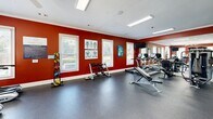 Located at 3606 Kaliste Saloom Rd in Lafayette, Louisiana, this dedicated fitness center offers a fully outfitted exercise environment designed to support a wide range of workout routines. The space is defined by bold, deep-red walls accented by crisp white crown molding and trim, creating an energetic and motivating atmosphere. The durable rubber flooring provides a safe and practical surface suited for both cardio and strength training activities. Multiple windows fitted with blinds allow ample natural light to fill the room, contributing to an open and airy feel. A large wall of floor-to-ceiling mirrors spans one side of the space, visually expanding the room while allowing users to monitor their form during workouts. The equipment selection is comprehensive, including multiple treadmills, an elliptical, and a stationary bike for cardiovascular training, alongside a cable resistance machine, a weight bench, a dumbbell rack, and medicine balls for strength and functional fitness. A wall-mounted flat-screen television is positioned for visibility from the cardio equipment. Recessed and linear fluorescent lighting ensures consistent illumination throughout. Anatomical and instructional fitness posters adorn the walls, adding both an educational and decorative element. Access is provided through elegant French-style glass doors, completing the polished presentation of this well-appointed fitness facility.