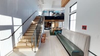 This two-story space showcases a contemporary open-plan design that emphasizes volume and connectivity between levels. The main floor features light-toned hardwood flooring that runs throughout, creating a cohesive and airy foundation. A long, sleek green leather bench lines one wall, providing ample seating while adding a bold accent to the otherwise neutral palette. A dining area anchors the central space, furnished with a wooden table and coordinating teal chairs atop a green area rug, introducing a playful pop of color. The staircase, constructed with natural wood treads and industrial-style metal railings, ascends along the left wall and serves as both a functional and architectural feature. Tall, multi-pane windows along the right wall flood the interior with abundant natural light, casting warm patterns across the floor. Above, a mezzanine level overlooks the main floor, offering an open railing view and additional functional space. The double-height ceiling in the main area enhances the sense of openness. The overall design blends industrial elements with warm natural materials, creating a dynamic and welcoming environment.