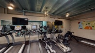 Located at 405 Broadway St in Cincinnati's downtown, this fitness center offers a well-equipped, single-floor workout facility with a distinctly industrial aesthetic. The space features exposed structural beams and ductwork painted in teal, complemented by stylish pendant and chandelier lighting that adds a contemporary touch to the raw, urban character of the room.

The cardio zone is outfitted with a strong lineup of equipment, including treadmills, elliptical trainers, and a rowing machine by BodyCraft, all positioned in front of large wall-mounted mirrors and flat-screen televisions for an engaging workout experience. Rubber flooring throughout provides both durability and comfort underfoot.

The strength training area houses a variety of resistance machines with blue padded seating, a cable weight station, and additional benches, catering to a full range of muscle groups. Wall-mounted bicycles add a creative use of vertical space and hint at cycling options as well.

A dedicated functional fitness corner rounds out the facility, stocked with stability balls in multiple sizes, resistance bands, exercise mats, and medicine balls neatly organized on a rack. Colorful wall art adds a vibrant, motivating energy to this area. A compact beverage refrigerator provides added convenience for hydration. Overall, the space balances industrial character with practical, comprehensive fitness amenities.