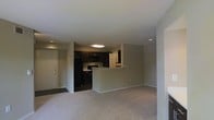 This single-floor space is designed with an open-concept layout that connects the living area and kitchen into one cohesive environment. The living area features neutral-toned carpet flooring and light gray walls, creating a clean and understated aesthetic that lends itself well to a variety of furnishing styles.

The kitchen is positioned toward the rear of the space and is distinguished by dark espresso cabinetry that provides a striking contrast against the lighter surroundings. A peninsula-style counter with a raised breakfast bar serves as a natural divider between the kitchen and living area, offering both functional workspace and casual seating potential. The kitchen is equipped with black appliances, including a refrigerator, microwave, and range, and is finished with light-toned countertops. A lighter upper cabinet section near a doorway provides additional storage.

Recessed lighting and a central flush-mount ceiling fixture illuminate the space effectively. A white entry door with a paneled design is visible to the left, providing a clear sense of entry and flow. The overall ambiance is modern, neutral, and move-in ready.