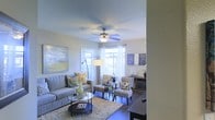 This single-floor space offers a comfortable and stylish living environment centered around a beautifully designed living area. The room is bathed in natural light streaming through large windows dressed with sheer white curtains, creating an airy and welcoming atmosphere. A ceiling fan with an integrated light fixture serves as a practical and decorative focal point overhead, helping to maintain a comfortable environment year-round. The living area is thoughtfully arranged with a plush gray sofa, complemented by patterned accent chairs and a sleek glass coffee table resting atop a textured area rug. Decorative touches, including bold wall art and carefully chosen accessories, add personality and warmth to the space. An arc floor lamp provides additional ambient lighting, enhancing the room's inviting ambiance. Dark hardwood-style flooring grounds the space and contrasts elegantly with the light-toned walls and soft furnishings. A media console along one wall offers functional storage and display space. The overall design strikes a balance between modern sensibility and comfortable livability, making this a versatile and welcoming space suited for both relaxing and entertaining.
